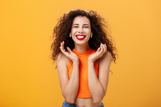 Stylish happy attractive urban female with curly hairstyle. and nose ring smiling broadly feeling delighted listening favorite track in wireless earbuds raising palms near face posing over orange wall.