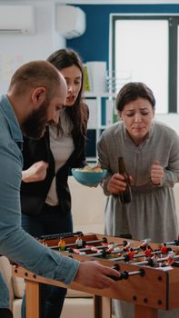 Woman winning game of soccer at foosball table while playing with man at office, having fun after work. Multi ethnic colleagues playing for entertainment, enjoying beer drinks.