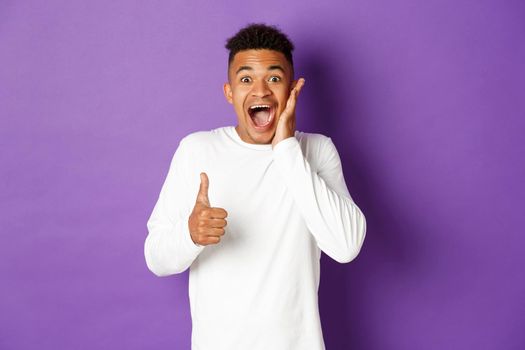 Portrait of amazed african-american man, showing thumbs-up and looking excited, recommend and praise good product, standing over purple background.