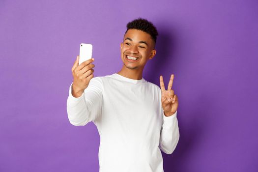 Cheerful african-american male blogger, taking selfie on smartphone, showing peace sign and smiling at mobile phone camera, standing over purple background.