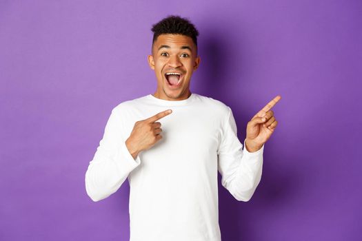 Portrait of handsome african-american guy in white sweatshirt, pointing fingers at upper right corner and smiling amazed, pointing fingers right at something cool, standing over purple background.
