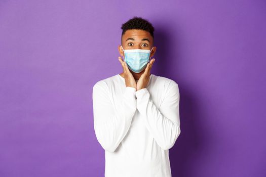 Concept of covid-19, pandemic and social distancing. Surprised african-american man, wearing medical mask and looking amazed, standing over purple background.