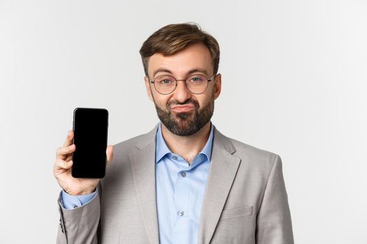 Close-up of skeptical businessman in glasses and gray suit, pouting and showing mobile phone screen, standing over white background.
