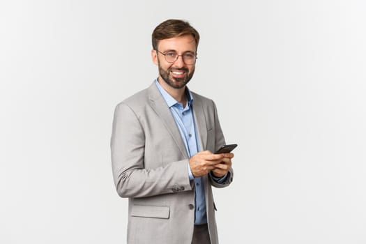 Handsome businessman with beard, wearing glasses and suit, holding mobile phone and smiling at camera, standing over white background.