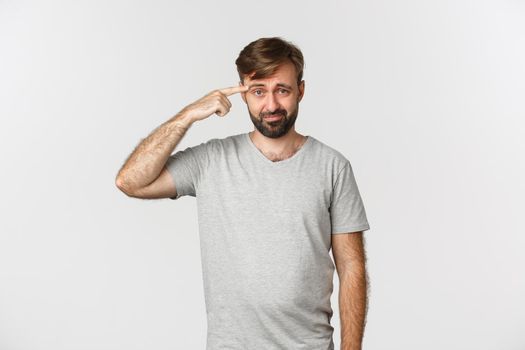 Portrait of frustrated guy scolding someone for being crazy or strange, pointing at head and frowning, standing over white background.