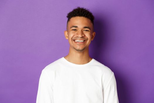 Close-up of young african-american man, smiling pleased, standing satisfied over purple background, wearing white sweatshirt.