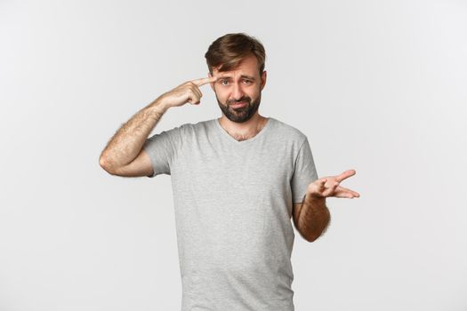 Portrait of frustrated guy scolding someone for being crazy or strange, pointing at head and frowning, standing over white background.