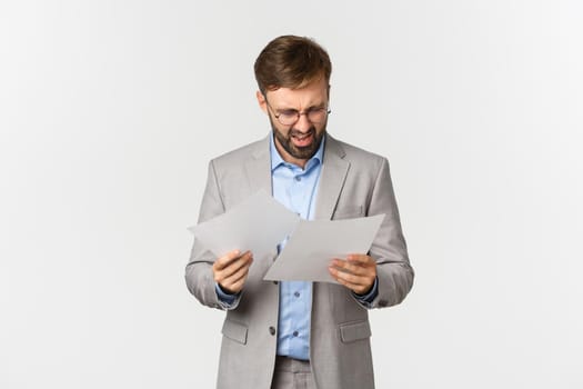 Portrait of angry and confused businessman in grey suit, looking at documents or bad report, standing disappointed over white background.