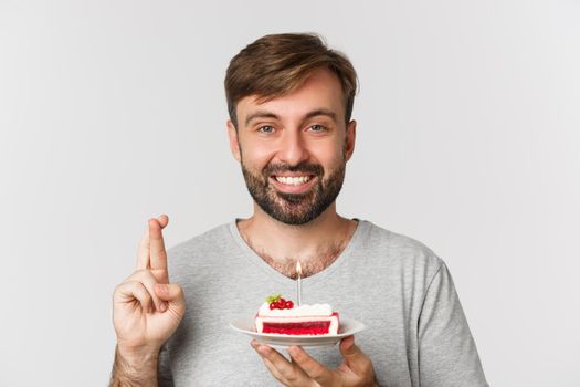 Close-up of hopeful man celebrating birthday, making wish with fingers crossed, holding bday cake, standing over white background.