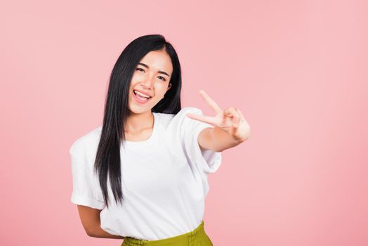 Asian happy portrait beautiful cute young woman teen smile standing show finger making v-sign victory sign gesture side away looking to camera studio shot isolated on pink background with copy space