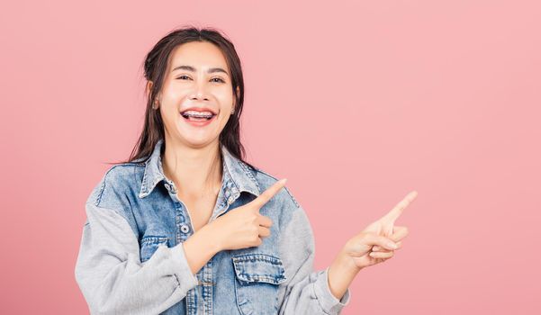 Asian happy portrait beautiful cute young woman wear denim standing pointing finger side away presenting product looking to camera, studio shot isolated on pink background with copy space