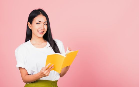 Portrait of beautiful Asian young woman teenage smiling holding a yellow book and reading, studio shot isolated on pink background with copy space