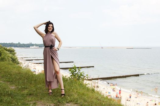 young brunette woman in beige dress standing by the sea on summer day