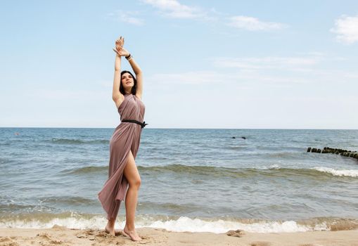 young brunette woman in beige dress stands on the sand by the sea on summer day