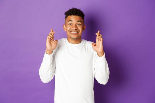 Portrait of hopeful african-american male model in white sweatshirt, cross fingers for good luck and looking at camera, smiling and making wish, standing over purple background.