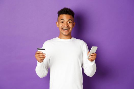 Portrait of handsome african-american man, smiling and looking excited while shopping online, holding credit card and mobile phone, standing over purple background.