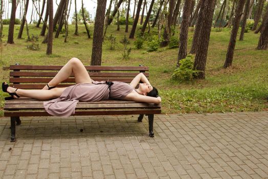 young beautiful brunette woman in beige dress lying on a park bench on summer day