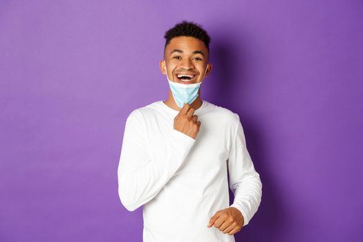 Concept of covid-19, pandemic and social distancing. Happy african-american man taking-off medical mask and breathing freely, standing over purple background.