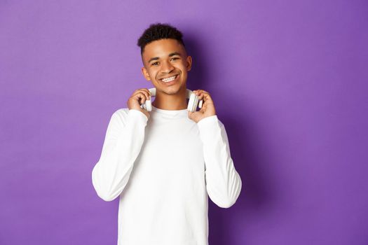 Image of cheerful african american man in white sweatshirt, holding headphones and smiling at camera, standing over purple background.