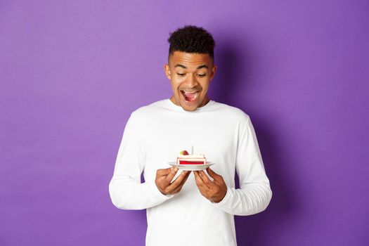 Portrait of happy young african-american man celebrating his birthday, looking at b-day cake and smiling, making wish, standing over purple background.