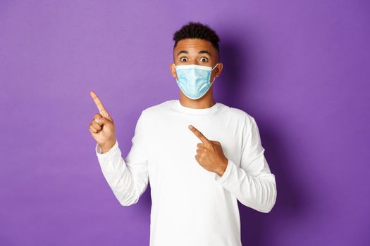 Concept of coronavirus, quarantine and lifestyle. Amazed african-american man in medical mask showing advertisement, pointing at upper left corner and smiling, standing over purple background.