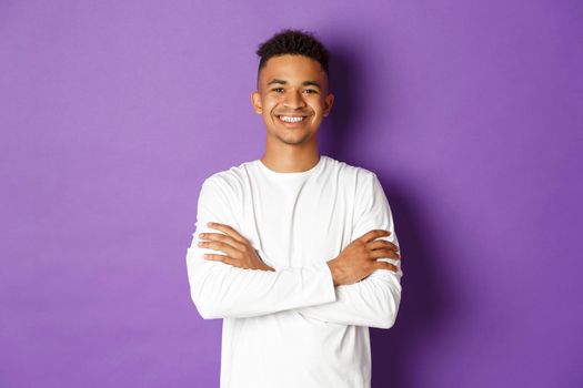 Portrait of handsome african-american male student in white sweatshirt, cross arms on chest and smiling confident, standing pleased over purple background.