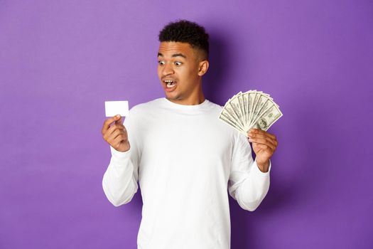 Image of handsome african american guy, showing money and looking excited at credit card, standing over purple background.