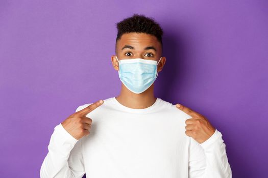 Concept of coronavirus, quarantine and social distancing. Close-up of young african-american man smiling, recommending to wear medical mask during covid-19, standing over purple background.