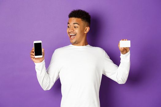 Portrait of handsome african-american man, looking amazed at mobile phone screen and showing credit card, standing over purple background.