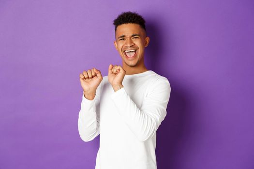 Image of silly african-american male model in white sweatshirt, dancing with satisfaction and pouting, celebrating achievement and victory, standing pleased over purple background.