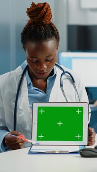 Woman doctor pointing at tablet with horizontal green screen, explaining technology to patient at desk. Medic holding chroma key with mockup template and isolated background on gadget.