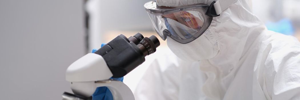 Scientist in protective suit and mask looks through microscope. Chemist in overalls working with various bacteria concept