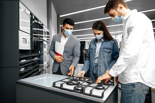 Shop assistant in hypermarket shows new model of gas stove to young couple, all wearing medical masks, close up