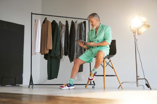 Full length shot of stylish middle aged man wearing colorful outfit using tablet pc while sitting next to clothes rail and studio spotlight over white background. Fashion photoshoot, style concept