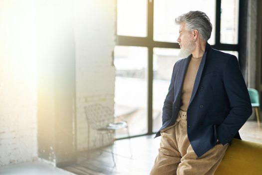 Stylish confident mature man looking aside while posing, leaning on sofa indoors. Lifestyle, people concept