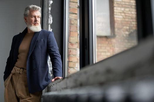 Pensive middle aged man in business casual wear looking out a large window, being thoughtful while standing in loft interior. Lifestyle, people concept