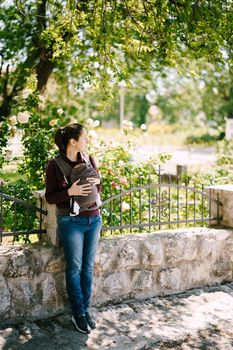 Mom with a baby in a sling stands leaning on a stone fence. High quality photo
