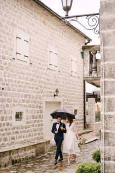 Bride and groom walk past the stone buildings on the cobblestones under an umbrella. High quality photo