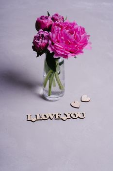Pink garden peonies on a gray textured background. Wooden inscription I love you. Valentine's Day greeting card. Postcard declaration of love. Wooden decor. Vertical image orientation.