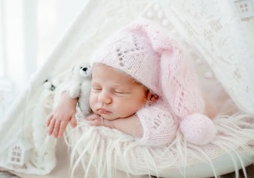 Newborn baby girl holding knitted teddy bear toy and sleeping on fur wearing hat. Infant female child kid napping pink color studio portrait