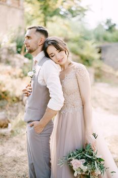Bride with a bouquet in her hand hugs groom from behind with her head on his shoulder. High quality photo