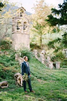 Groom with a bouquet stands near the ancient chapel in the garden. High quality photo