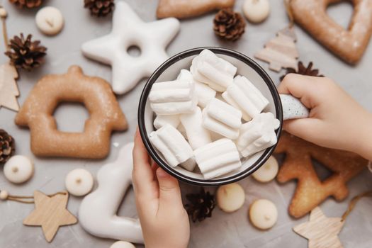 Children's hands hold a cup of cocoa with marshmallows. Christmas gingerbread on the table, bokeh lights in the foreground. The concept of desserts and drinks during the Christmas holidays.