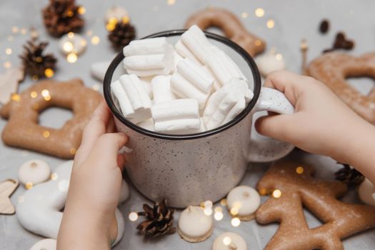 Children's hands hold a cup of cocoa with marshmallows. Christmas gingerbread on the table, bokeh lights in the foreground. The concept of desserts and drinks during the Christmas holidays.