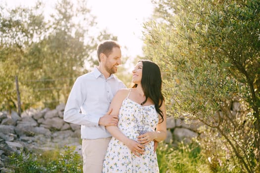 Man hugging from behind a pregnant woman holding wildflowers. High quality photo