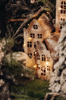 Stock photo of handmade cardboard Christmas buildings on green lawn and surrounded by green Christmas tree branches decorated with garland.