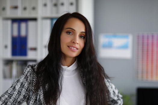 Portrait of attractive young businesswoman wearing stylish jacket posing in company office. Brunette corporate worker smiling. Business, career concept
