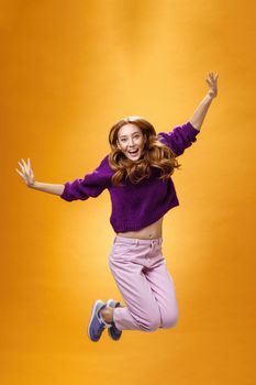 Expressive and enthusiastic carefree friendly joyful redhead woman in purple sweater and pants jumping joyfully with raised hands smiling yelling from happiness and joy against orange background. Copy space