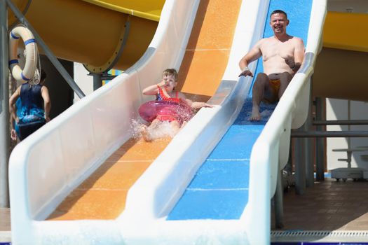 Portrait of father and daughter slide down hill on air mattress in water park. Smiling parent and child enjoy summer holiday. Summertime, leisure concept