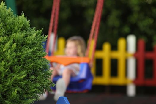 Close-up of green fluffy bush on playground child on swing in park alone. Little girl enjoy ride outdoors, place for children. Childhood, nature concept